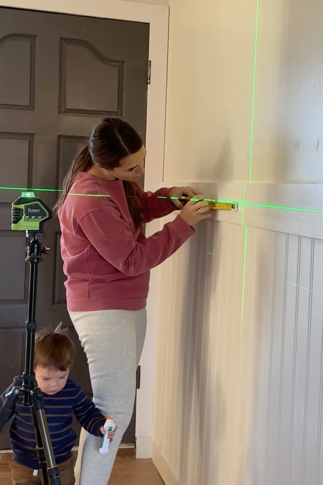A woman using a measuring tape and level, guided by bright green laser lines, to install a wooden peg rail onto a freshly installed beadboard panel. A toddler holding a toy watches nearby, adding a family touch to the DIY process.